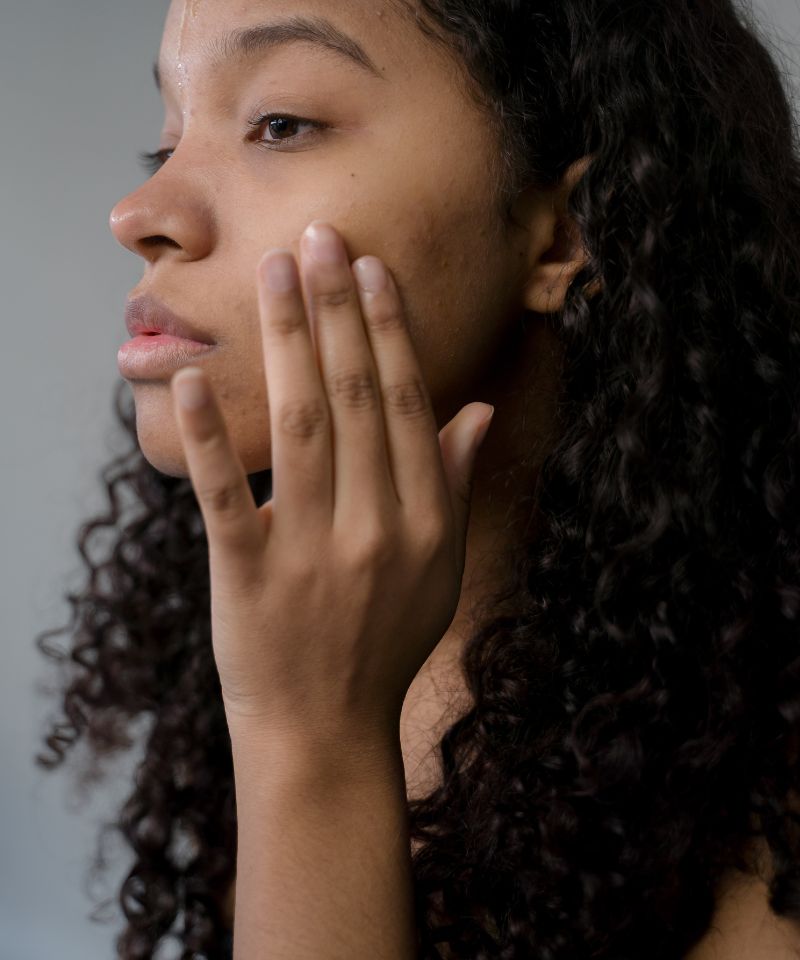 A dark skinned female with a worried expression on her face looking at her face in the mirror trying to spot signs of acne rosacea.
