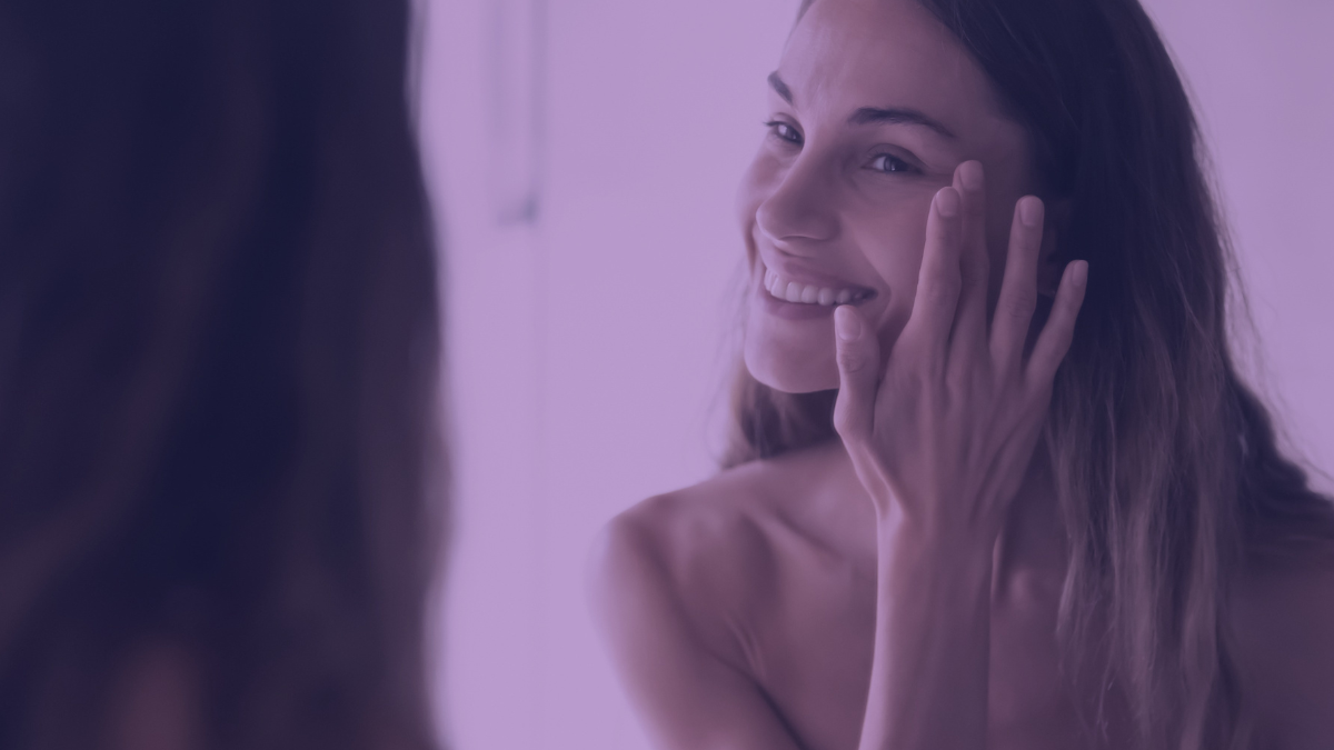 A woman smiling in the mirror as she applies her skincare routine.