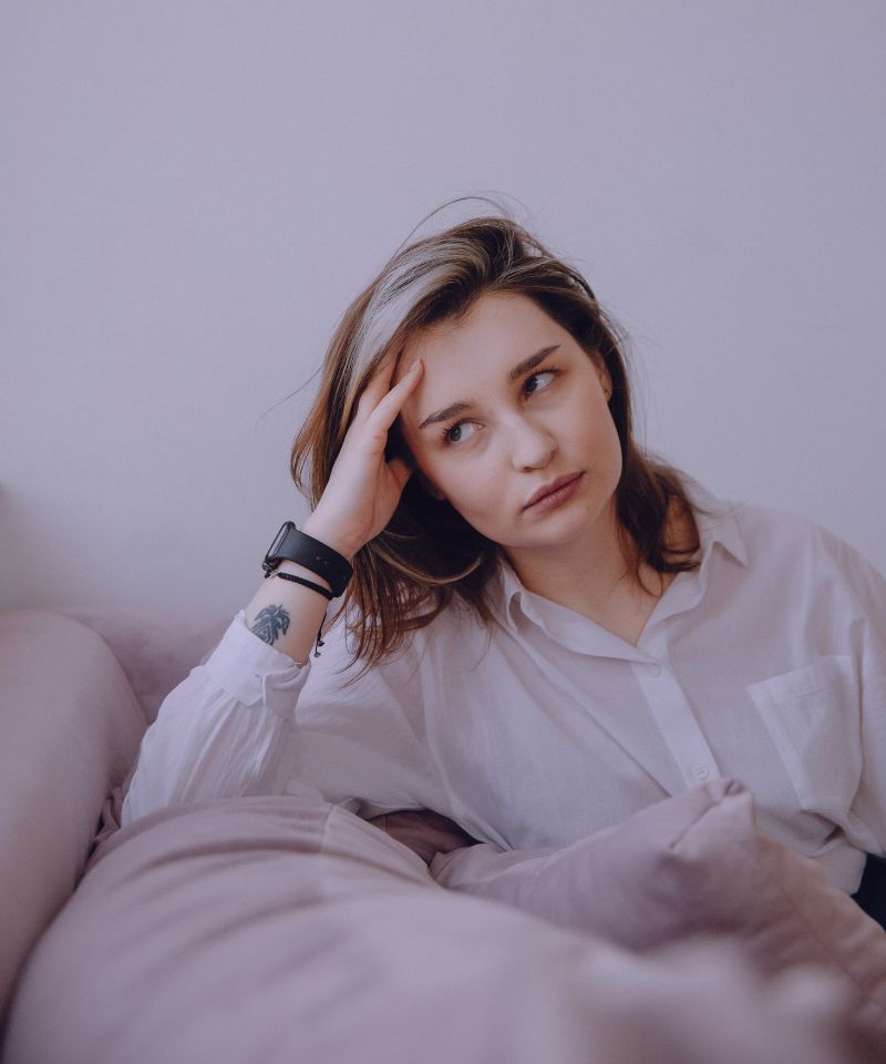 A pensive young person sits, leaning on a cushioned hand, slightly turned away from the camera. Managing stress is vital for skin health, as it can ward off stress-induced skin issues like acne. Mindfulness, exercise, and rest are key in controlling stress, helping to preserve skin health. The individual's calm setting and posture may represent the positive effects of pausing to unwind and reflect, a beneficial stress management strategy.