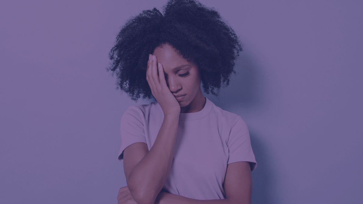 This image depicts a person appearing to be stressed or in discomfort. They have their eyes closed and are pressing their forehead with their fingers, suggesting a headache or a moment of frustration. The individual has a medium skin tone and their hair is styled in a natural afro, indicating that they might be of African descent. The person is wearing a simple, plain t-shirt and the background is a calm, uniform purple, emphasizing the subject without distraction. Stress can have various effects on the skin, including the exacerbation of acne. When stressed, the body produces more of the hormone cortisol, which can lead to an increase in oil production in the skin glands. This excess oil can clog pores, leading to the development or worsening of acne. Additionally, stress might also affect the skin's barrier function, potentially making it more sensitive and reactive, further contributing to skin issues like acne.